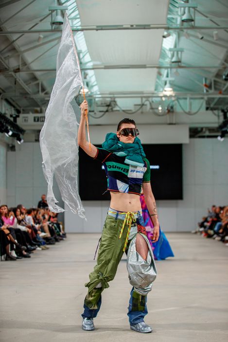 Person on runway wearing green pants with silver patch, cropped top, and holding a large white flag.