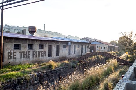 Langes Gebäude mit flachem Dach und der Aufschrift "BE THE RIVER" an der Seite, davor ein mit Gras bewachsener Graben.
