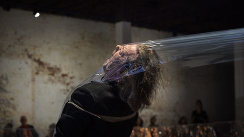 Person mit langem Haar, die den Kopf nach hinten neigt und von einer durchsichtigen Folie umhüllt ist, vor unscharfem Hintergrund mit mehreren Personen.