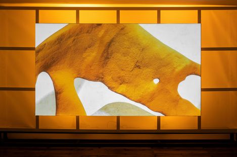 Wide screen showing a yellow-orange abstract form against a glowing background in a gallery space.