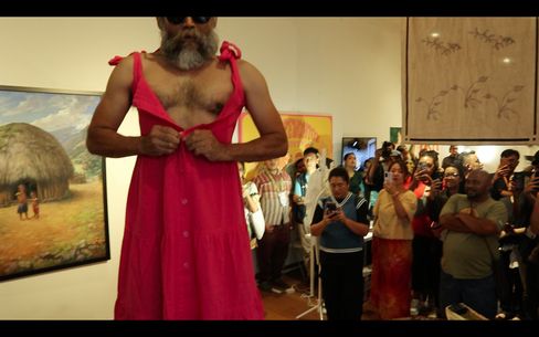 Mann mit rotem ärmellosem Kleid, der vor einer Gruppe von Menschen in einem Raum mit Kunstwerken steht