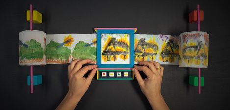 Hands holding a long painted paper strip with repeated yellow and black patterns attached to two rods with colorful handles.