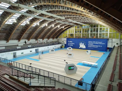 Large sports hall with curved wooden roof structure and multiple fencing pistes on the floor, a large blue banner with text and a map of Europe on one wall.