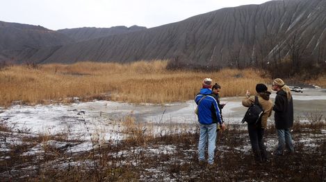 Four people stand on a partially frozen, marshy area in front of a steep, eroded slope with bare bushes.