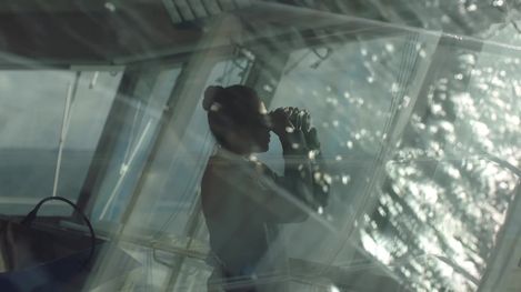 Silhouette einer Person mit zusammengebundenem Haar, die durch ein Fernglas auf das Wasser blickt, aufgenommen durch ein Fenster mit Reflexionen.