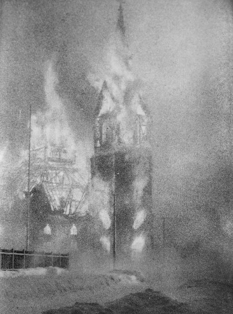 Church tower and building engulfed in flames and smoke in a historical black-and-white photo.
