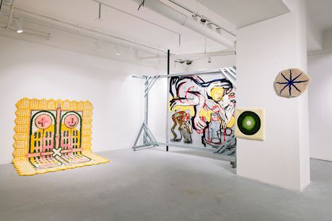 Exhibition space with several abstract artworks on walls and floor including a yellow patterned piece on the floor, a large wall painting with human figures, and two round greenish and beige artworks on the wall.