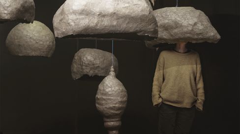 Person standing behind a large irregularly shaped paper-like sculpture covering their head, surrounded by other similar sculptures of various shapes and sizes.