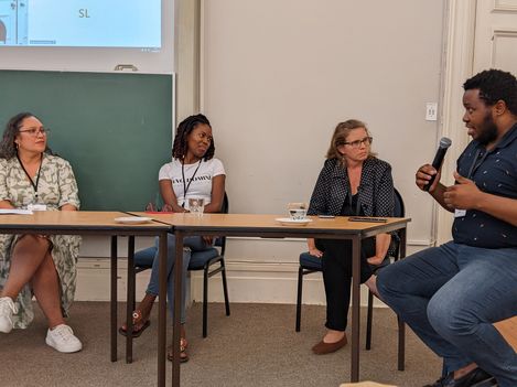 Four people are sitting at a table in a room with a blackboard and a projection screen. The person on the right side of the image is holding a microphone. A discussion is taking place.