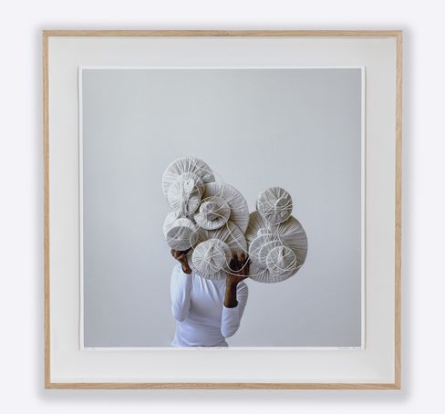 Person wearing a white top holding several round objects wrapped in white yarn in front of their face.