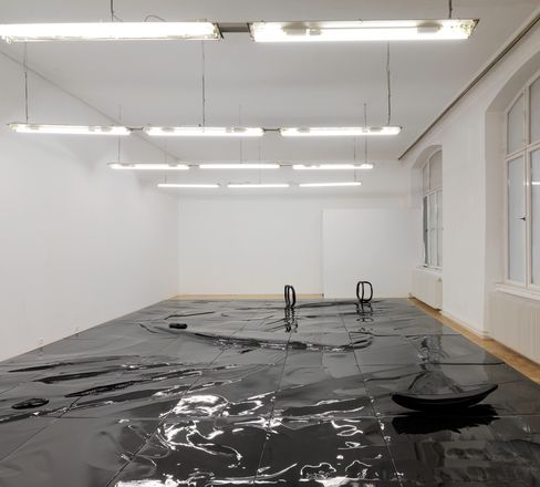 Large room with white walls and ceiling lights, shiny black sheet on the floor with two black handles and a black round object.