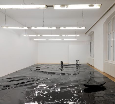 Large room with white walls and ceiling lights, shiny black sheet on the floor with two black handles and a black round object.