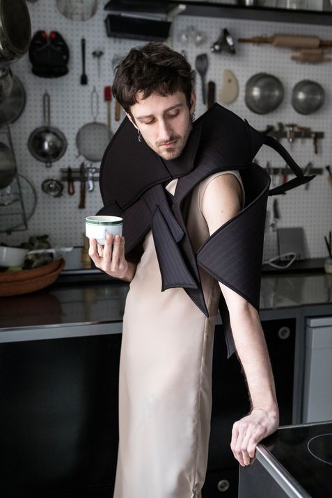 Person with short hair wearing a beige dress and a black sculptural garment with wide padded elements in a kitchen with hanging cooking utensils.