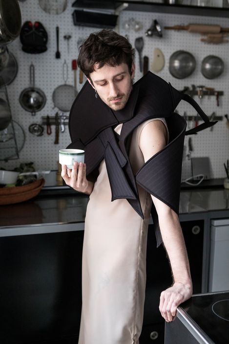 Person with short hair wearing a beige dress and a black sculptural garment with wide padded elements in a kitchen with hanging cooking utensils.
