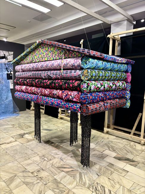 Sculpture made of stacked colorful fabric rolls on a raised structure in a gallery.
