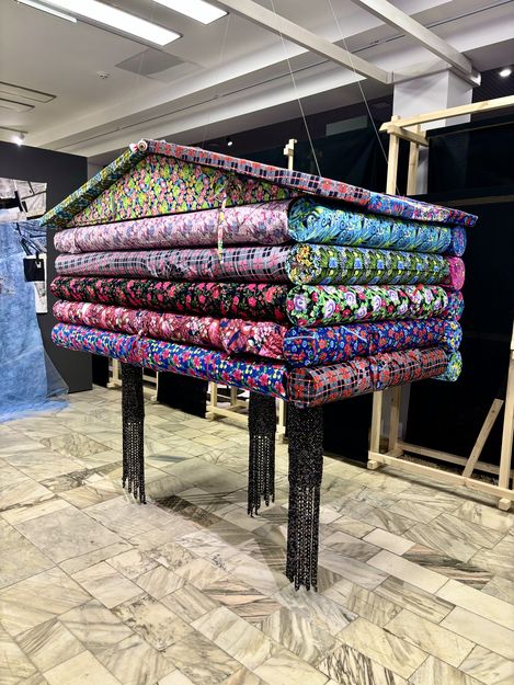 Sculpture made of stacked colorful fabric rolls on a raised structure in a gallery.
