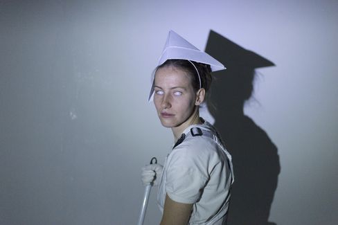 Person wearing a paper hat and white T-shirt standing sideways against a wall with shadow.