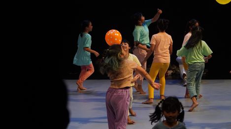 Mehrere Kinder in bunter Kleidung spielen mit einem orangefarbenen Ball auf einer Bühne mit dunklem Hintergrund.