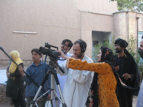 Group of people with a camera and microphone outdoors in front of a light-colored wall.
