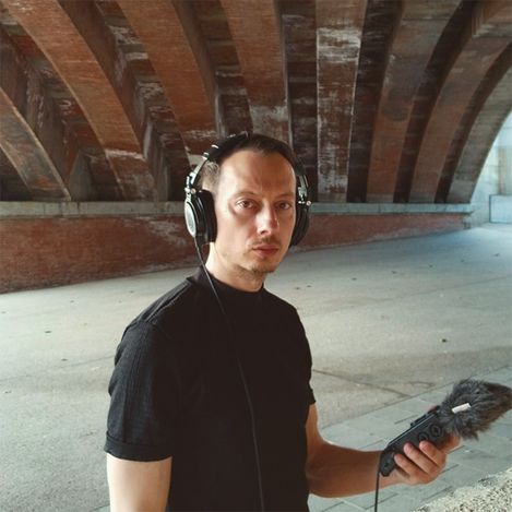 Person mit Kopfhörern und Mikrofon unter einer Brücke mit sichtbaren Betonbalken.