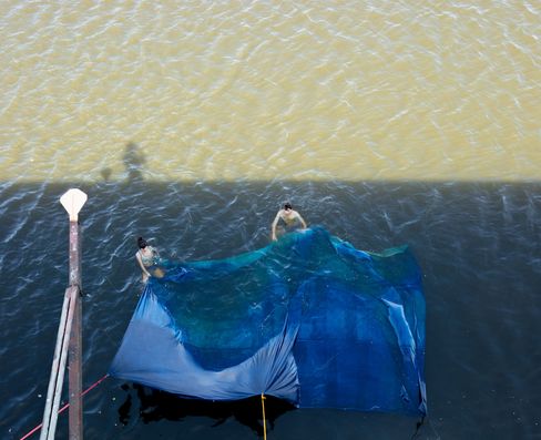 Two people in water holding a large blue net next to a pier.