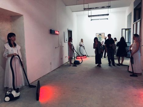Mehrere Personen in einem weißen Raum, einige tragen weiße Kleider und halten Staubsauger, eine rote LED-Anzeige an der Wand zeigt "U bahn 16 4".