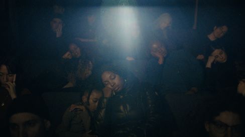 In a dark cinema, several people sit with their eyes closed beneath a bright beam of light shining down from above.