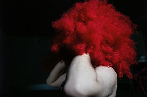 Person with voluminous curly red wig against dark background holding their head with both hands.