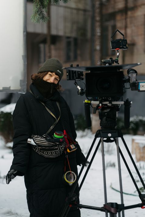 Person in Winterkleidung steht neben einer professionellen Filmkamera auf einem Stativ im Freien.