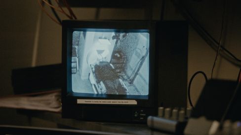 A small monitor displays a grainy black-and-white image in a room filled with cables and technical equipment.