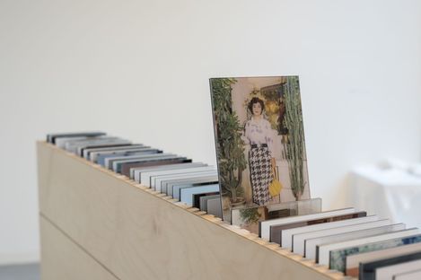 Holzregal mit mehreren aufrecht stehenden Postkarten, eine Postkarte zeigt eine Frau in gemustertem Rock und gelber Tasche vor Pflanzen.