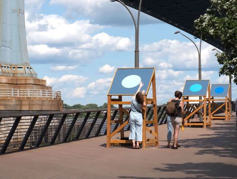 Zwei Personen betrachten große Tafeln mit kreisförmigen Motiven auf einer Brücke unter blauem Himmel mit Wolken.