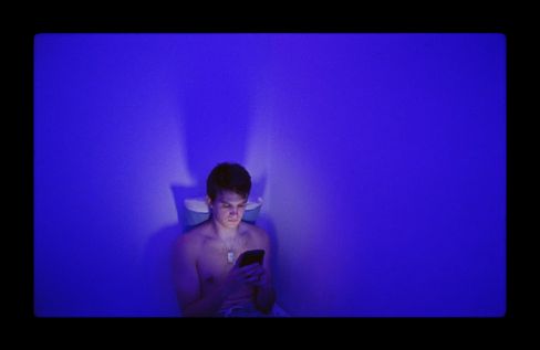 A young person sits shirtless on a toilet bathed in blue light, looking at a smartphone.