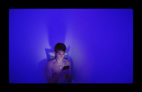 A young person sits shirtless on a toilet bathed in blue light, looking at a smartphone.