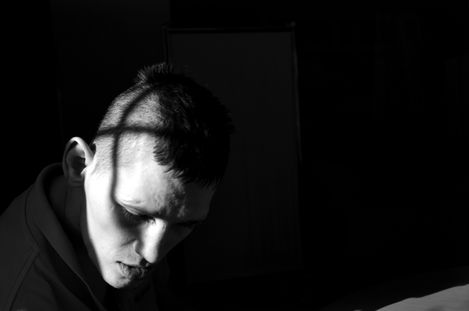 Portrait of a young man with short hair looking downward with a grid shadow on his face against a dark background.