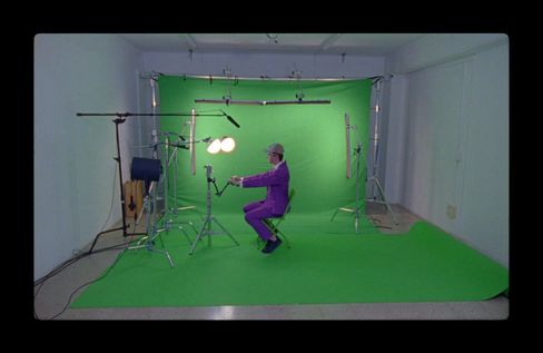 A person wearing a purple suit sits in front of a green screen in a film studio with visible lighting and microphones.