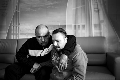 Two men sit close together on a sofa, the older man embracing the younger, both looking downward.