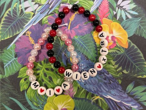 Nahaufnahme von zwei bunten Perlenarmbändern mit Buchstabenperlen, die „MIA“ und „WIENXTRA“ ergeben, arrangiert auf einem farbenfrohen Hintergrund mit tropischem Muster aus Blättern und einem blauen Vogel.