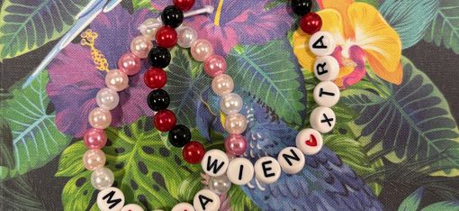 Nahaufnahme von zwei bunten Perlenarmbändern mit Buchstabenperlen, die „MIA“ und „WIENXTRA“ ergeben, arrangiert auf einem farbenfrohen Hintergrund mit tropischem Muster aus Blättern und einem blauen Vogel.