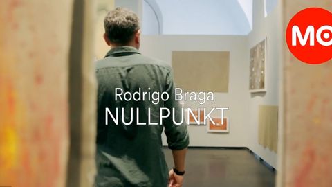 A man seen from behind in an exhibition space. A red logo with white lettering reading “MQ” in the corner and white text reading “Rodrigo Braga NULLPUNKT” in the center of the image.