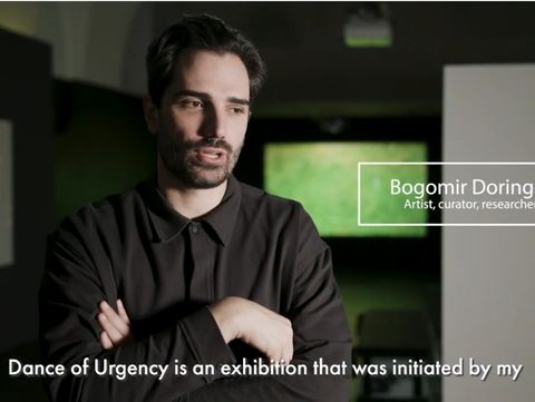 A person with crossed arms, wearing a dark shirt, speaks in a dimly lit room. Text identifies them as an artist, and captions mention an exhibition.