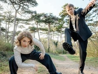 Two people in formal clothing pose on a forest path, one crouching and the other lifting a leg.