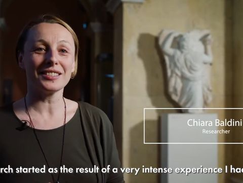 A woman smiles in a museum hallway with warm lighting. A marble statue is blurred in the background. Text reads: "Chiara Baldini, Researcher."
