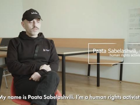 A person in a black hoodie and cap, sitting in a modern room with a wooden bench and tables, identified on-screen as a human rights activist from Georgia. The tone is calm and serious.