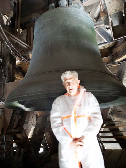 Ein Mann in weißem Schutzanzug steht vor einer großen Glocke in einem hölzernen Glockenturm.