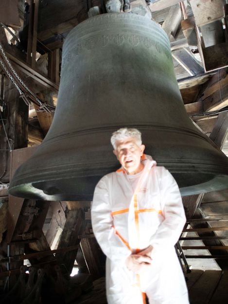 Ein Mann in weißem Schutzanzug steht vor einer großen Glocke in einem hölzernen Glockenturm.