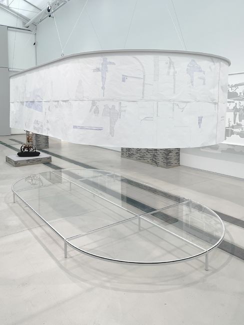 Großes, ovales, transparentes Tischgestell aus Metall und Glas in einem weißen Raum mit einem darüber hängenden, halbtransparenten, ovalen Stoffelement mit abstrakten Mustern.