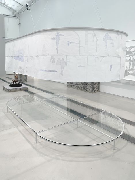 Großes, ovales, transparentes Tischgestell aus Metall und Glas in einem weißen Raum mit einem darüber hängenden, halbtransparenten, ovalen Stoffelement mit abstrakten Mustern.