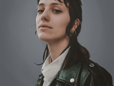Person with dark shoulder-length hair, multiple earrings, and black leather jacket against gray background.