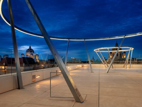 Dachterrasse mit großen, leuchtenden Metallringen auf geneigten Stützen vor einem abendlichen Himmel und historischen Gebäuden im Hintergrund.